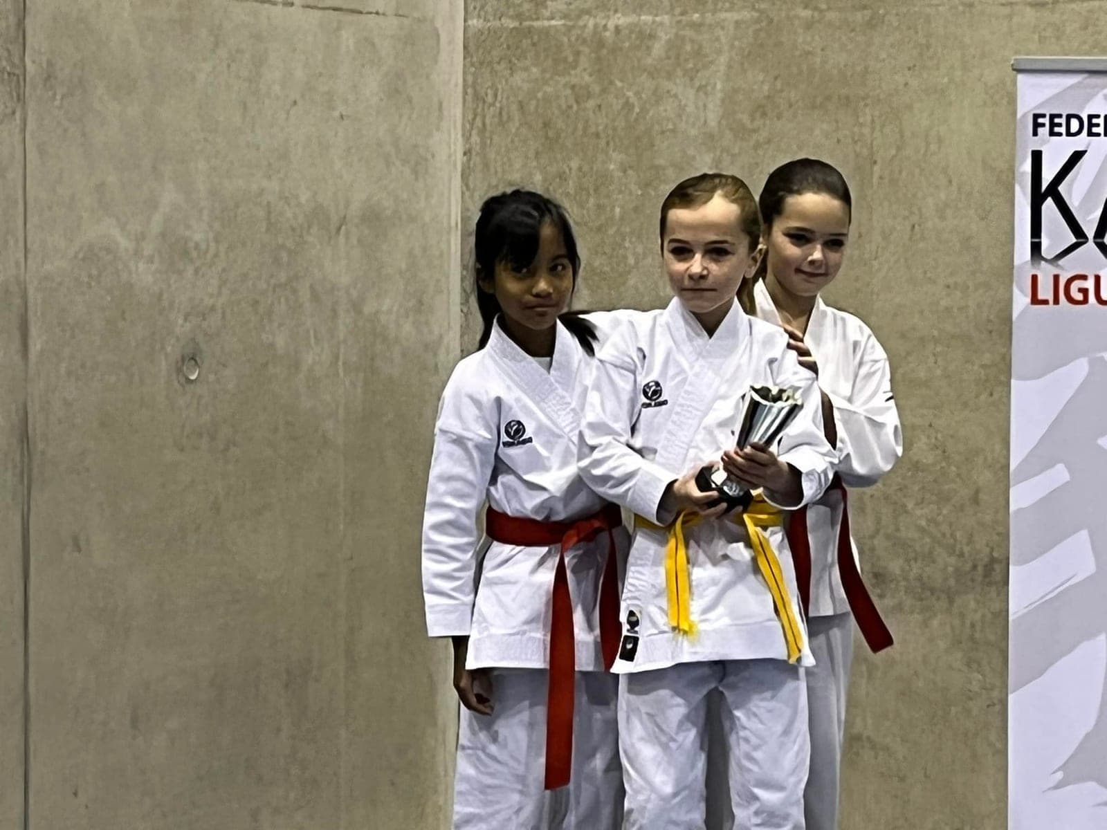 Trois jeunes karatékas sur le podium, l’une d’elles tenant la coupe, devant une banderole de la Fédération française de karaté