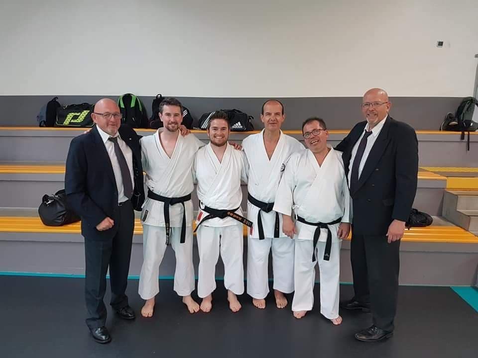 Quatre karatékas ceinture noire entourés de deux dirigeants en costume, réunis devant les gradins d’une salle de sport