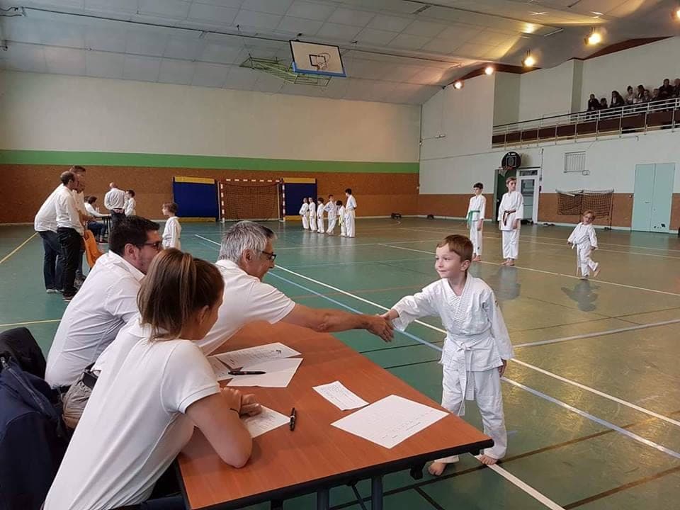 Un jeune karatéka en karategi blanc serre la main d’un membre du jury assis derrière une table d’examen, dans un gymnase où d’autres enfants attendent leur tour