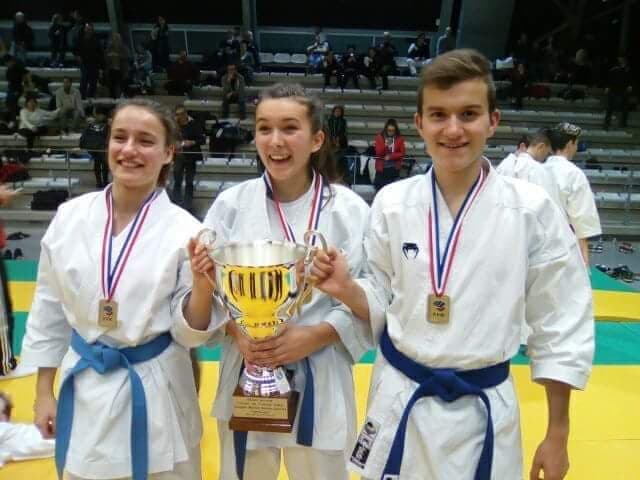 Trois jeunes compétiteurs en karategi, médaille autour du cou, tiennent ensemble une coupe dorée devant les gradins d’un gymnase